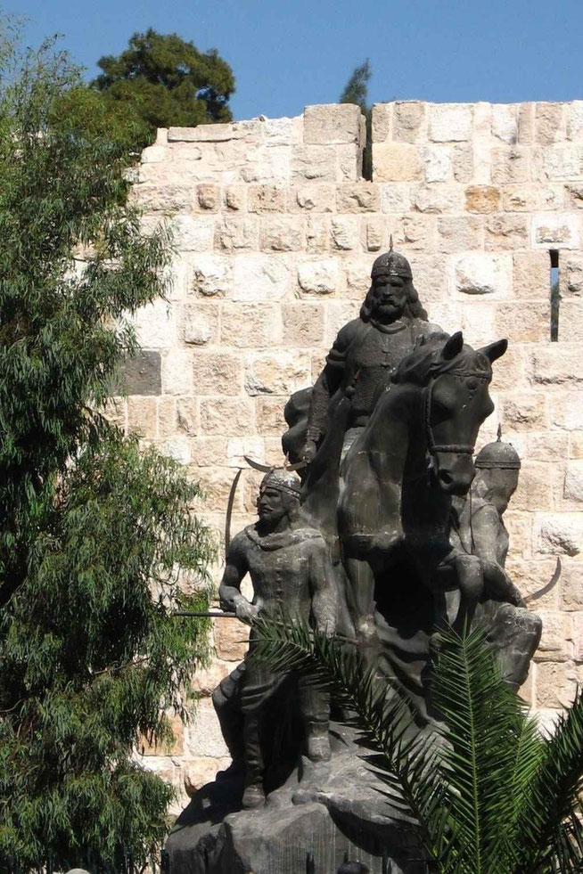 Bronze statue of a mounted warrior surrounded by armed figures, set against a stone wall with trees and clear blue sky.