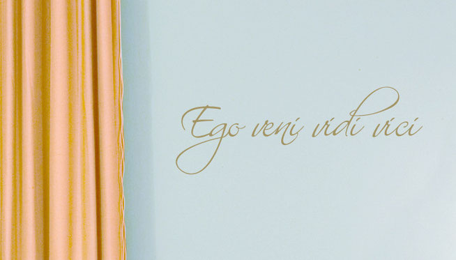 Ego Veni Vidi Vici - I came, I saw, I conquered, metallic gold wall art sticker in an elegant handwriting font, on a light blue wall near gold curtains in a living room.