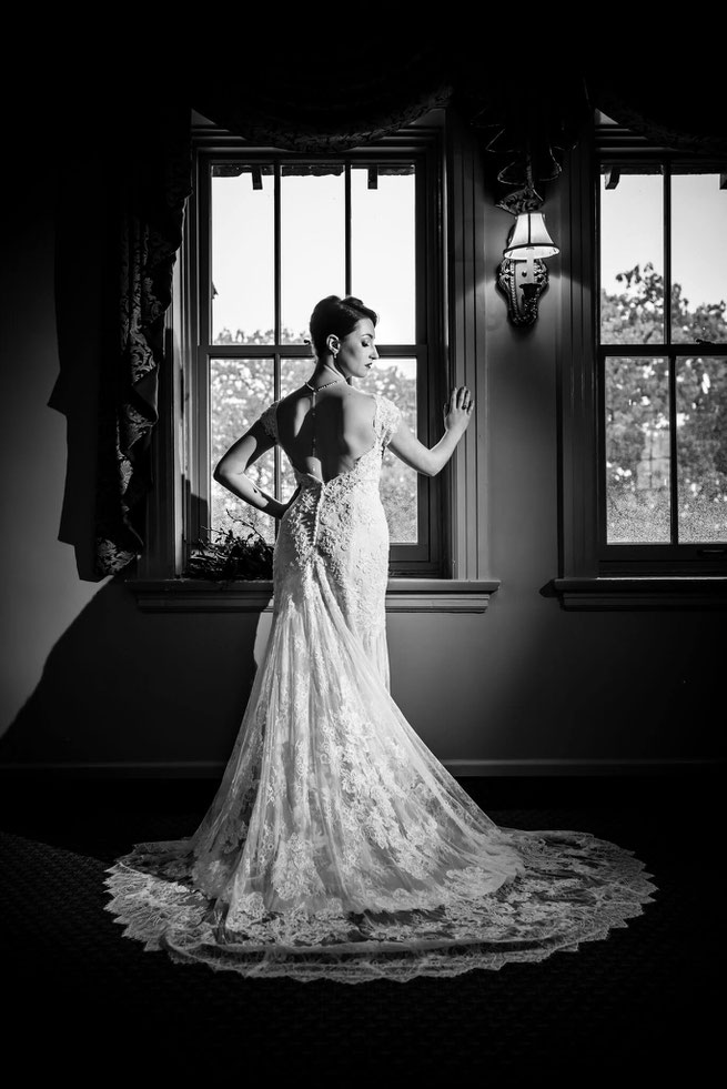 Black and white photo of a bride in front a window. Bride is shown from behind with face to the side. Dress is fanned on floor. 