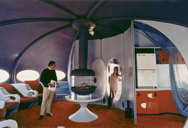 Interior de una Futuro, mostrando parte de la sala con una novedosa chimenea redonda, la puerta de la recámara (por donde sale una mujer) y parte de la cocina