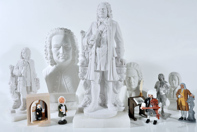 Etwa 15 Bach-Büsten und Bach-Figuren aus Gips, Plastik, Holz und Zinn sind zu einem Gruppenbild vor weißem Hintergrund und auf weoißem Untergrund arrangiert.