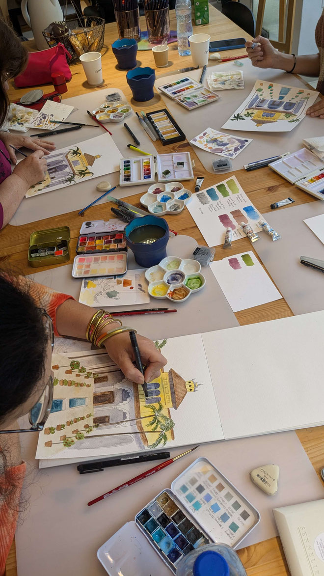 Une table avec des outils de peinture à l'aquarelle, incluant des pinceaux, des crayons, des palettes de couleurs et des tubes de peinture. Des échantillons de couleurs et des illustrations de bâtiments sont présents.