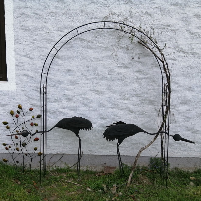 Zwei schwarze Metall-Vogelskulpturen, die sich voneinander abwenden, unter einem Rankbogen vor einer weißen Hauswand.