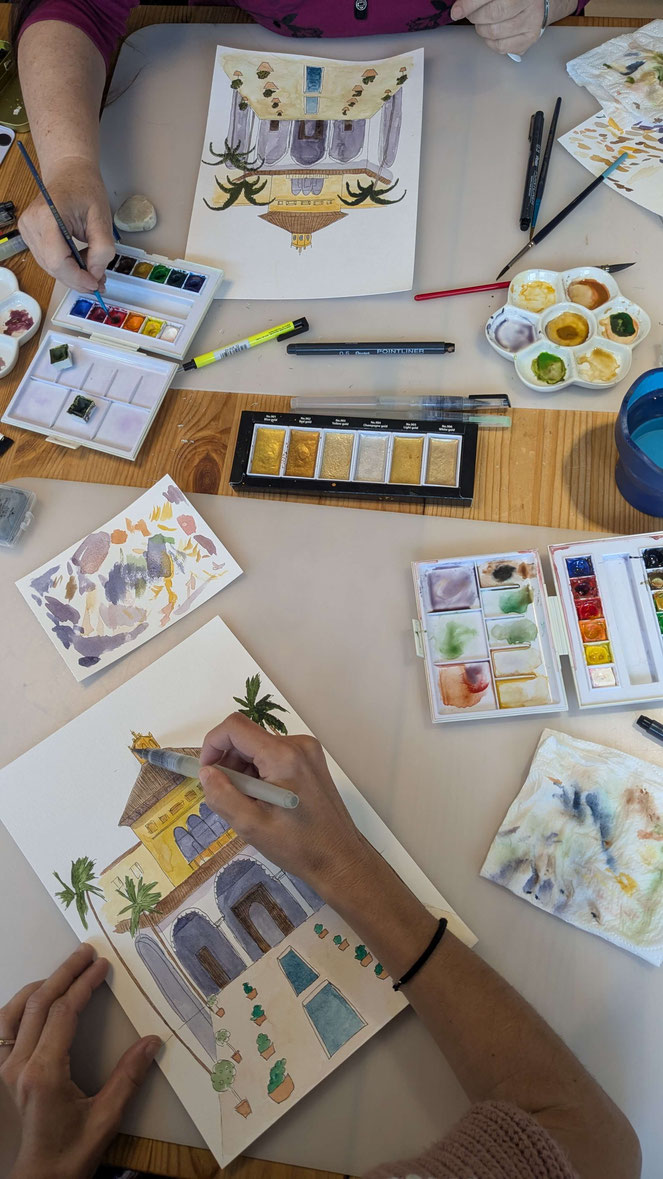 Deux personnes peignent à l'aquarelle avec des pinceaux et des crayons. La table contient des palettes de couleurs, des tubes de peinture et des illustrations de bâtiments avec des détails architecturaux et des plantes.