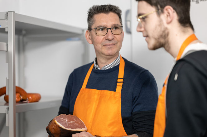 Ausbilder erläutert Azubi in der Kühlung die Lagerung eines Rohschinkens.