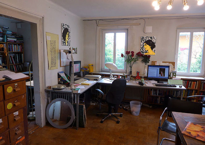 Großes Zimmer mit zwei Fenstern, Parkett, großem Eckschreibtisch und zwei PC-Bildschirmen, Magazinschrank aus Holz, an den Wänden Bilder von Virginia Woolf und Vita Sackville-West