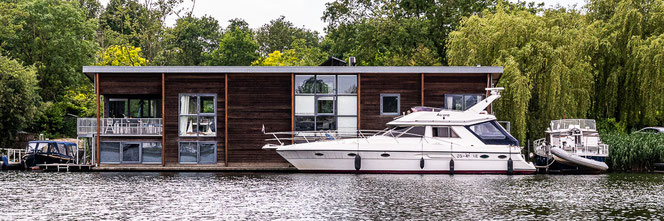 Ultramodernes schwimmendes Haus auf dem Wasser umgeben von mehreren Booten. Vor der langgestreckten Hausfront eine ultra-chice weiße Yacht.