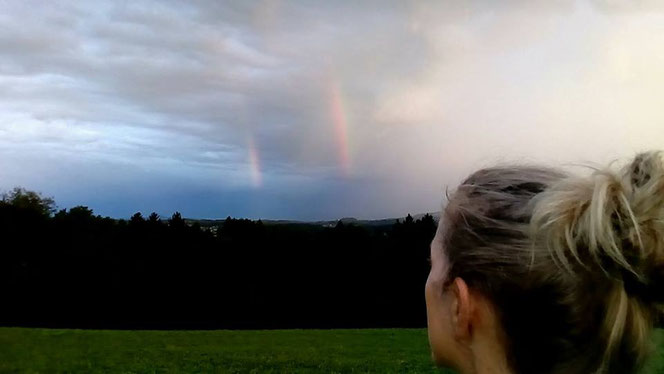 Christine Harnik blickt über eine Wiese in die Landschaft; am Himmel sind zwei schwache Regenbögen zu sehen.