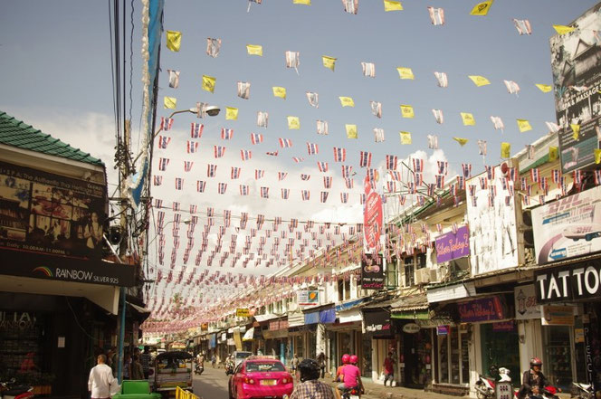 バックパッカーの聖地・バンコク カオサンエリア 数え切れないタイ国旗、タイ王室の旗などが翻る。写真の右側左側は商店。中央には道路。ピンク色のタクシー、ピンク色のバイクにまたがる人も映る【バンコク ピクチャー】  タイ王国の首都・バンコクの旅行（出張）写真ブログ