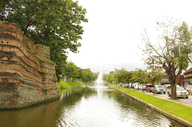 チェンマイの城壁とその手前の水堀の光景。奥には噴水があり、水しぶきを上げている。右側には道路が整備され自動車、ソンテウが通行している。お濠と道路の間には緑の芝生がある。[タイ・チェンマイ旅行（出張）写真ブログの画像]