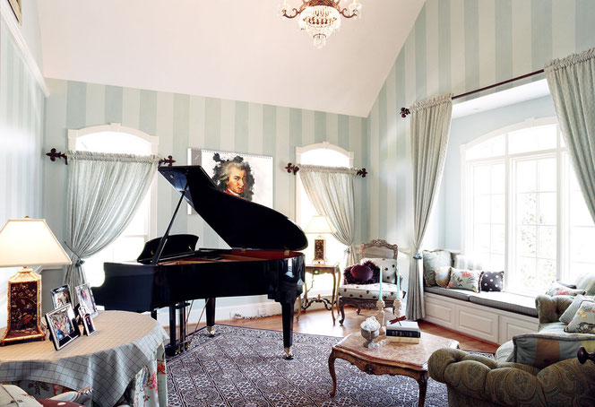 Eine Wohnzimmer mit Couchgarnitur und große Fenstern, der Ton ist hellgrün. Darin steht in der linken Hälfte ein schwarzer Flügel. An der Wand hinter dem Flügel hängt ein Komponisten-Kalender, man sieht Mozart.