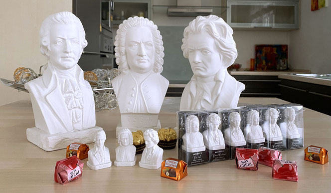 Auf einer Kücheninsel stehen in zweiter Reihe drei große Gipsbüsten von Mozart, Bach und Beethoven. In der vorderen Reihe sind 9 Radiergummis, links sind 3 ausgepackt. Außerdem sieht man verschiedene Süßigkeiten zum Größenvergleich.