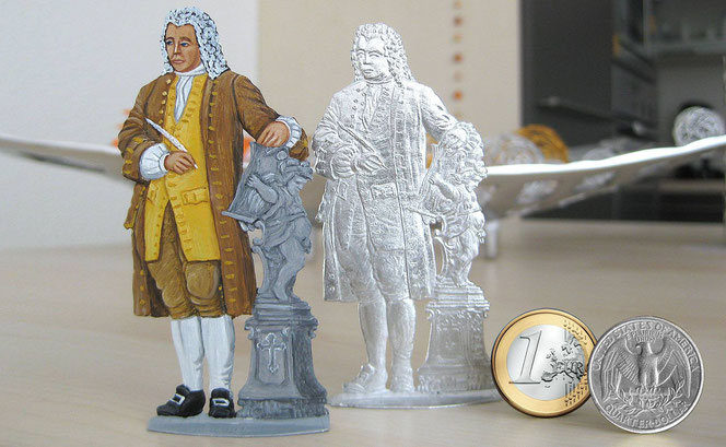 Zwei flache Bach-Zinnfiguren, eine bemalt, eine unbemalt stehen auf einer Kücheninsel vor einer Deko-Schale. Rechts sind außerdem zwei Münzen zum Größenvergleich: ein Euro und ein US-Quarter.