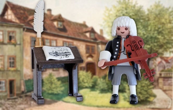 Das Playmobil-Bach-Männchen schwebt vor dem unscharfen Hintergrund, nämlich dem Bachhaus in Eisenach. Bach spielt Geige, links neben ihm sind Notenpult, Feder, Tintenfass und ein Notenheft.