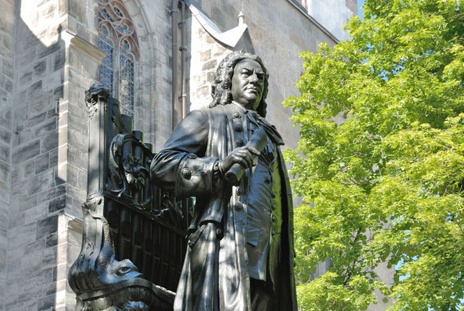 Ein Foto in Leipzig: Das Neue Bachdenkmal. Bach steht vor einer Orgel und hält gerollte Notenblätter in der Hand. Hinter Bach ist die Seite der Thomaskirche. Rechts neben Bach ist ein Baum im Frühlingslaub.