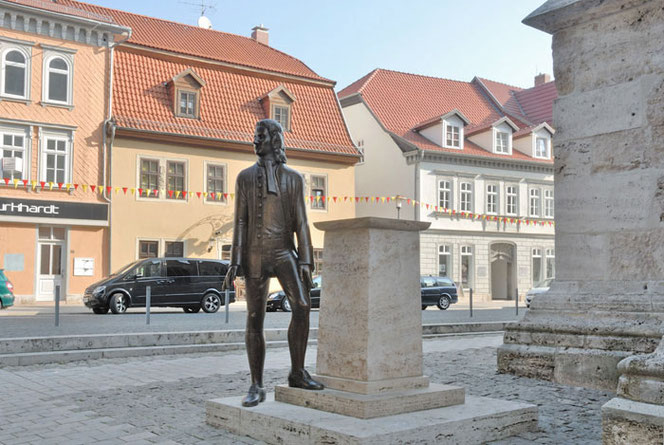 Bach in Mühlhausen. In der Innenstadt steht Bach neben seinem Podest neben einer Kirche. Er ist noch sehr jung. Das Denkmal ist nur etwa zwei Meter groß. Im Hintergrund ist eine Straße, Pkw und Häuser.