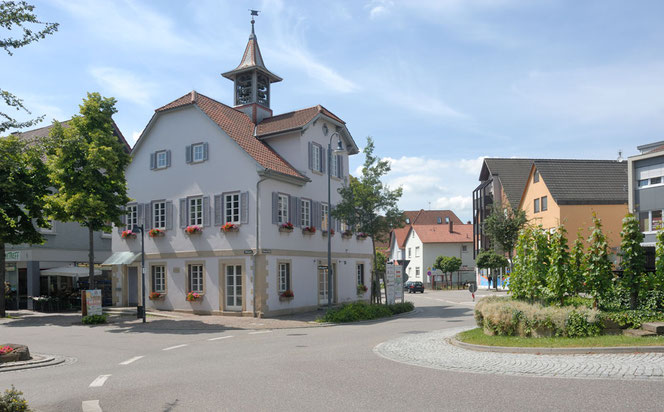 Links das Alte Rathaus in der Ortsmitte von Flein bei Heilbronn. Um das Rathaus herum führt eine Straße, die sich zwischen Häusern in der Tiefe des Bildes verliert. Rechts ist die Pflanzinsel in einem Kreisverkerhr. Man sieht keine Autos.