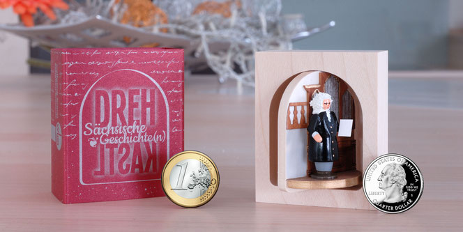 Auf einer Kücheninsel steht rechts das Bach-Drehkast'l mit Bach vor einer Orgel auf einem halbrunden Podest. Link sieht man die rote dekorative Schachtel dazu. Zusätzlich sieht man zwei Münzen, einen € und einen US-Quarter für einen Vergleich.