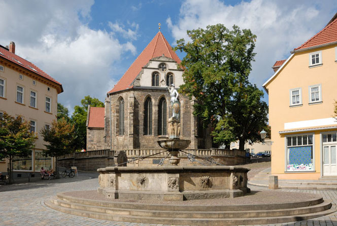 Der Blick zur Bachkirche in Arnstadt auf einer kleinen Anhöhe im Ort. Davor ist ein hübscher Brunnen. Die Kirche hat keinen Turm. Links und rechts sind Teile von Wohnhäusern. Es sind Wolken im blauen Himmel.