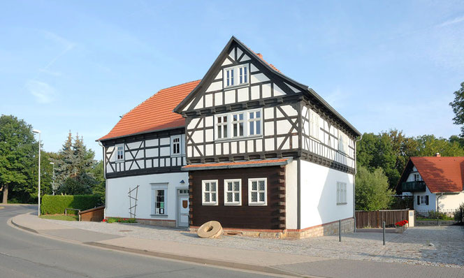 Es ist die Veit-Bach-Mühle in Wechmar bei Gotha bei bestem Wetter fotografiert vor blauem Himmel. Es ist ein Fachwerkbau, eine Straße umrundet die Mühle.