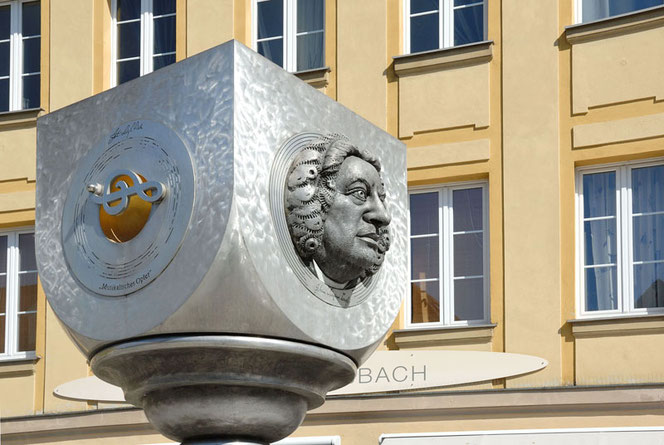 Das moderne Bach-Denkmal in Ansbach. Es ist ein silbern-metallener Kelch-Quader. Rechts an einer Seite ist das plastische Porträt von J.S. Bach, links ist ein Notenschlüssel auf einer kleinen goldenen Fläche. Im Hintergrund ist ein gelbes Geschäftshaus.