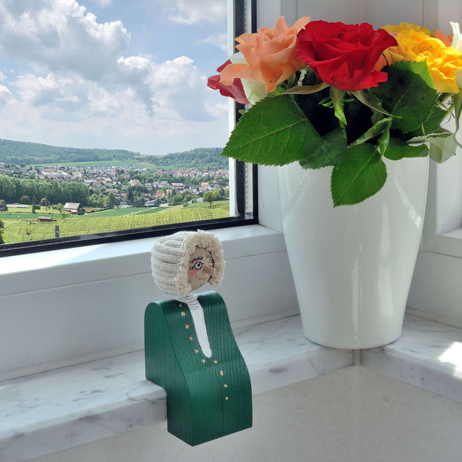 Der Bach-Kantenhocker sitzt auf einer weißen Fensterbank vor einem weißen Fenster. Im Hintergrund sieht man Weinberge, einen  Ort und Wald. Rechts neben Bach ist eine weiße Vase mit Blumen und Blättern.