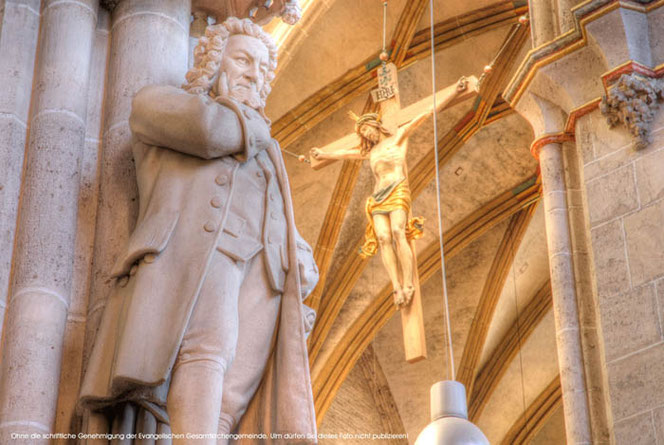 Im Ulmer Münster steht eine riesige Bach-Statue auf einem Sockel. Man blicjt steil nach oben. Im HIntergrund sieth man das Kreuz mit Christus. Der Hintergrund ist in sanften Gelbtönen. Bach ist in Marmor-Beige.