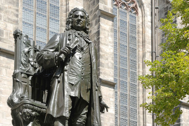 Das Johann-Sebastian-Bach-Denkmal in Leipzig. Es ist das neue von beiden. Im Hintergrund sieht man das Seitenschiff dr Thomaskirche. Rechts am Rand ist ein Baum mit Frühjahrslaub.