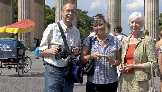 Drei Personen stehen vor den Säulen des Brandenburger Tors und lächeln herzlich in die Kamera. Es sind von links Peter Bach jr., Renate Bach und Petra-Ines Kaune. Links im Hintergrund ist ein Fahrzeug mit Pedalen und rot/gelbem Sonnendach.