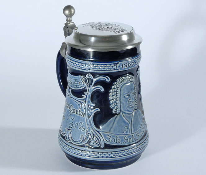 Der blaue Bach-Bierkrug aus dem Jahr 1985. Der Untergrund ist dunkelblau, die Designs sind hellblau. Oben ist ein einfacher Zinndeckel mit dem Siegel von Bach, hinten ist ein dunkelblauer Griff.