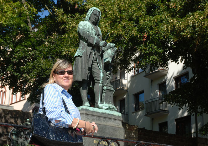 Renate Bach vor dem Bach-Denkmal in Eisenach. Sie lächelt herzlich in die Kamera, hat eine dunkelblaue Tasche umhängen und trägt eine Sonnenbrille. Hinter dem Bach-Denkmal sind Bäume. Das Motiv ist steil nach oben aufgenommen.