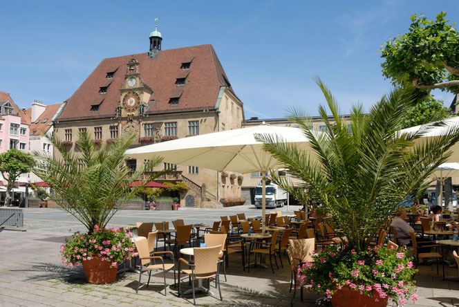 Im Hintergrund links oben ist das Heilbronner Rathaus. Ganz vorne rechts im Bild sind ausgebreitete Sonnenschirme eines Retsaurants mit Palmen und Bestuhlung.