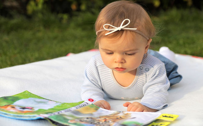 Ein niedliches kleines Mädchen schaut sich Bilder in einer Illustrierten an. Es hat ein hellblaues Outfit und liegt auf einer weißen Decke im Gras. Im Haar hat es eine süße Schleife.