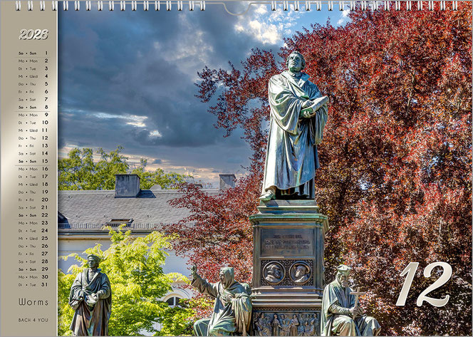 Ein Foto von einem Luther-Denkmal. Rechts ist bildfüllend ein Baum mit roströtlichem Sommerlaub. Unten sieht man weitere drei Figuren. Es ist ein Monatsblatt im Luther-Kalender. Links ist auf goldenem Untergrund das Kalendarium.