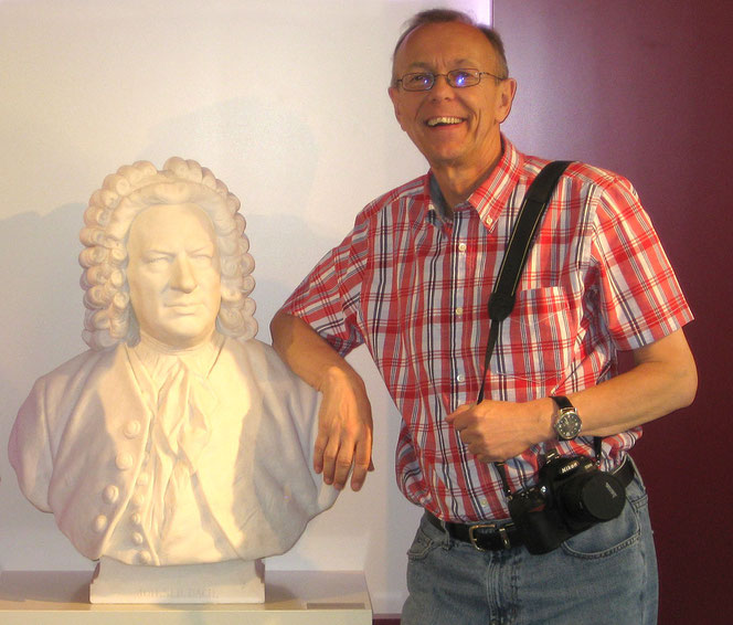 Peter Bach jr. (rechts) steht neben einer Johann-Sebastian-Bach-Büste (links). Er lehnt sich auf die Schulter der Büste auf. Im Hintergrund ist eine weiße-rote Wand zu sehen. Peter Bach jr. hat eine Kamera über der Schulter hängen.