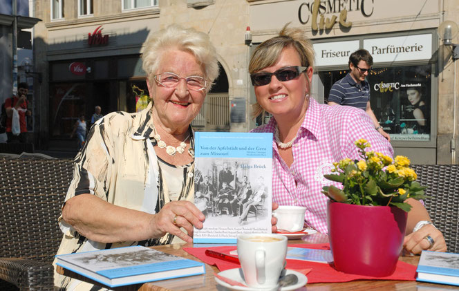 Zwei Damen sitzen in einem Café an einem Tisch. Die linke ältere Dame hält ein Buch in die Kamera. Auf dem Tisch liegen noch zwei solche Bücher. Beide lächeln herzlich zum Betrachter. Vorne sind ein roter Blumentopf und eine weiße Tasse mit Unterteller.