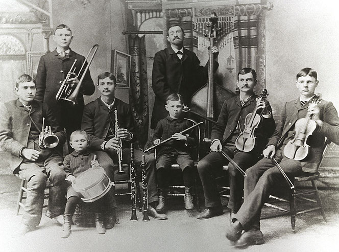 Das Gruppenbild einer historischen Band in den USA. Es ist die Bach-Band. Sie schauen alle zur Kamera und halten dabei ihre Instrumente. Es sind acht Personen, die Aufnahme ist schwarzweiß.