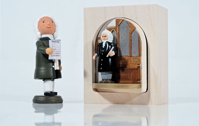 Auf einem weißen Untergrund und weißen Hintergrund stehten zwei Bach-Geschenkartikel. Links ist es ein kleines Bach-Männchen aus Holz, rechts ist es Bach in einem Drehkast'l vor einer Orgel. Beide Figuren sind winzig.