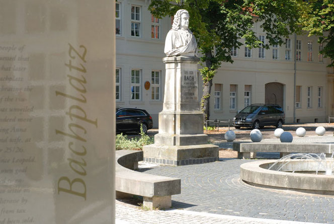 Das Bach-Denkmal in Weiß in Köthen. Im linken Bilddrittel ist riesig ein hübsches Hinweisschild mit dem Wort Bachplatz, in der Mitte ist das Denkmal, Bach auf vier Podeststufen. Rechts ist ein Springbrunnen. Im Hintergrund ist eine Hausfront.