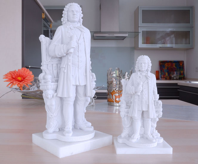 Zwei Bachstatuen stehen auf einer Kücheninsel nebeneiander, im Hintergrund ist die Küche. Beide Statuen sind aus Gips und weiß. Sie stehen nebeneinander und sind verschieden hoch. Bach steht vor einer Orgel und hält gerollte Notenblätter.