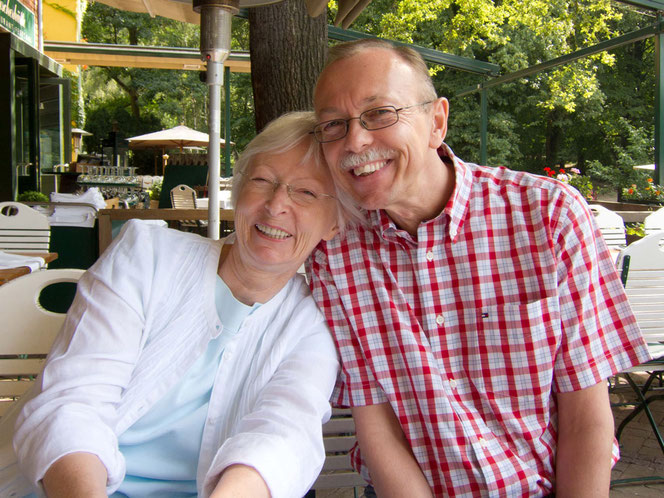 Peter Bach jr. und seine Schwester, Ines-Petra Kaune, lehnen in einem Restaurant im Außenbereich herzlich nebeneinander gegeneinander. Beide lächeln herzlich zum Fotografen. Sie hat ein weißes Outfit an, er ein kurzärmeliges rot-kariertes Hemd.