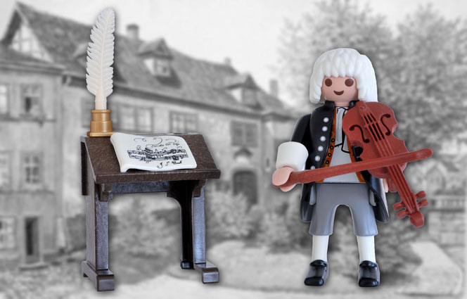 Die Kleine Bach-Figur schwebt vor dem unscharfen Hintergrund, dem Bachhaus in Eisenach. Rechts ist Bach, der auf einer Geige spielt. Links ist ein Notenpult mit Federkiel, Tintenfass und Notenheft.