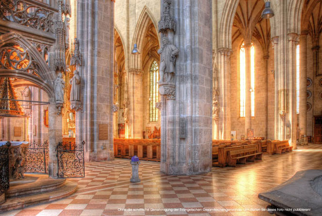 In the huge interior of Ulm Minster, you can see the Bach monument on a very high pedestal next to a column in the nave. The sun is shining into Ulm Minster, giving the whole picture a warm yellow color.