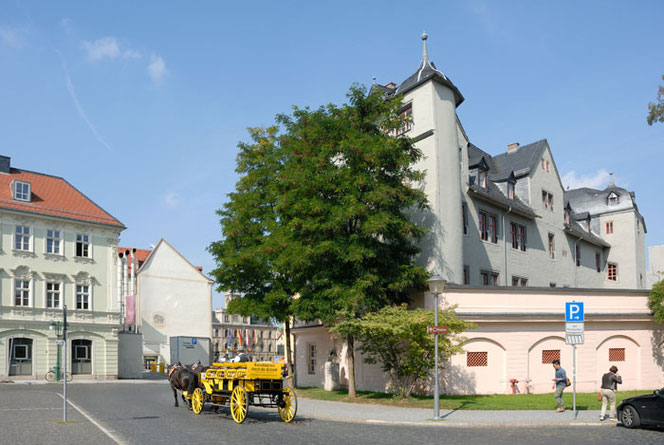 An ensemble of historic buildings with a yellow carriage in the lower center of the picture. The Bach memorial between two trees can be seen to the right of the carriage. In front is a paved road surface.