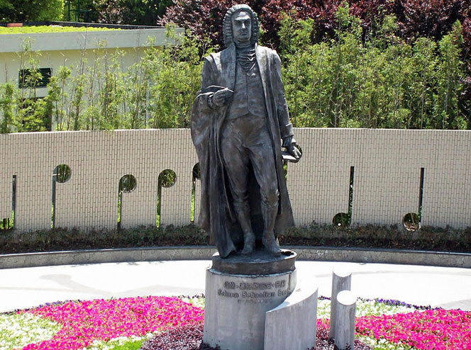 The Bach monument in Shanghai, China. It is very large, depicts Bach completely and standing, and is located in a green area. There are beautiful plants at the bottom. Johann Sebastian Bach is written on the pedestal in Chinese.