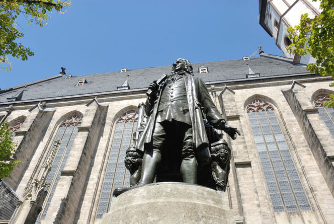 The New Bach Monument in Leipzig is photographed steeply upwards in front of the side of St. Thomas Church. Bach looks imposing. The pedestal is only slightly depicted, the sky is blue.