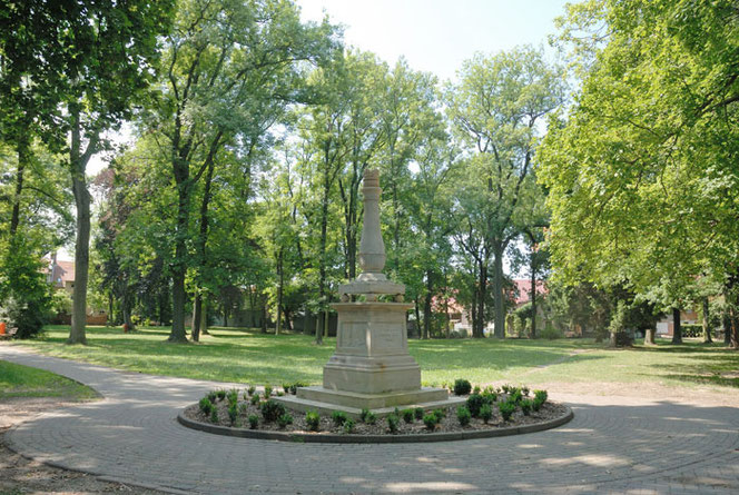 In a park with trees in spring there is a monument that consists only of a kind of obelisk. There are small plants in a circle around it. A path leads around it, which also goes down to the left.