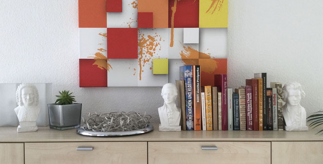 On a wall in an apartment you can see a sideboard surface with three gypsum busts of Bach, Beethoven and Mozart. Two busts serve as bookends for around 15 books. In the background on the wall is a comparatively large picture.