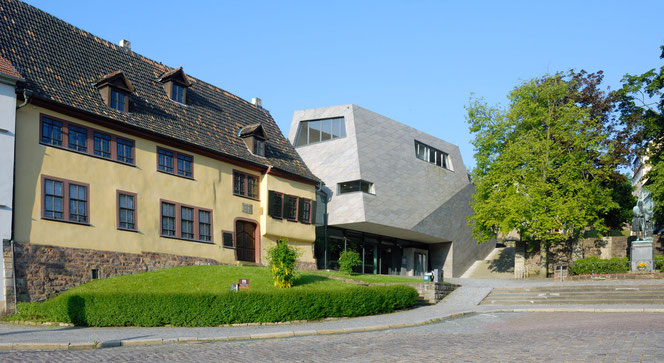 The Bach ensemble in Eisenach on the Frauenplan in the morning sun. On the left is the complete Bach House, in the middle is the super-modern Bach Museum, further to the right is a tree in spring foliage, to the right of that is the Bach Monument. The sky