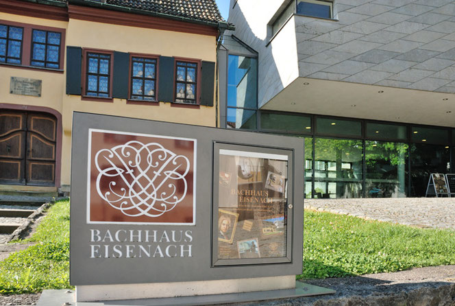 In front of the Bach House, half on the left and the Bach Museum on the right, there is an information board with the giant Bach Seal, albeit without a crown or laurel. There is a lawn between the sign and the buildings.